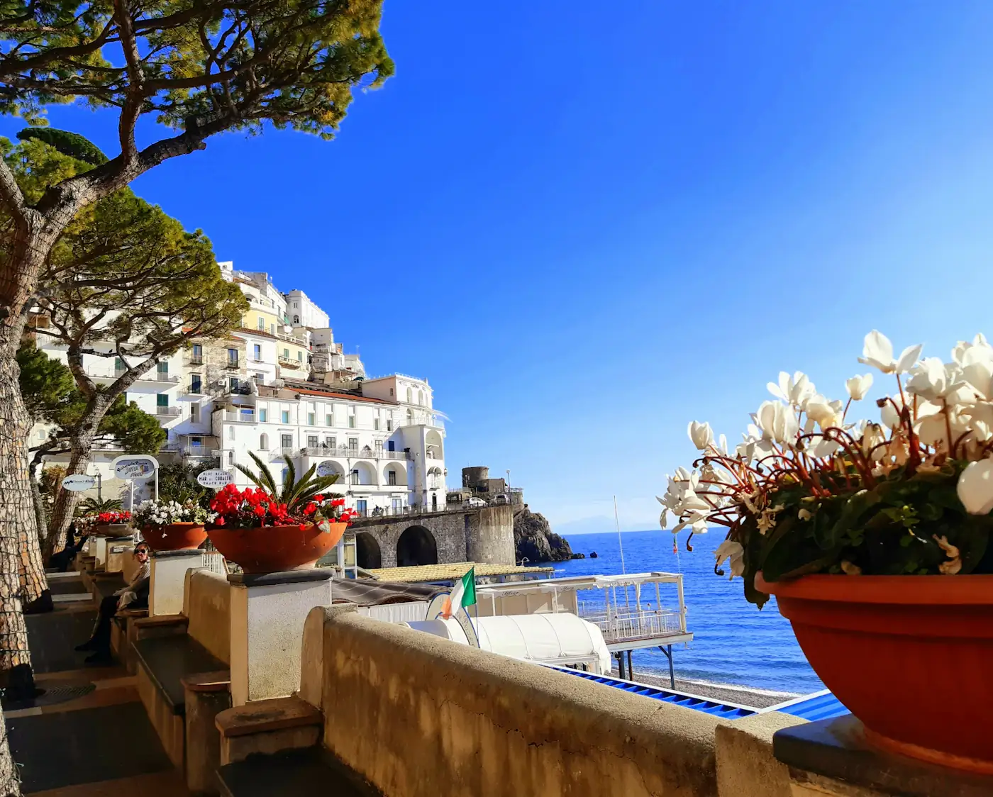 A terrace in Amalfi with potted flowers, white buildings and the sea behind.