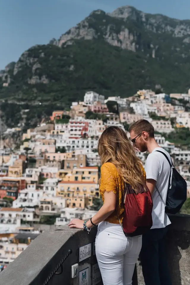 A couple at a railing looking out over Positano, its houses stacked up the hillside.
